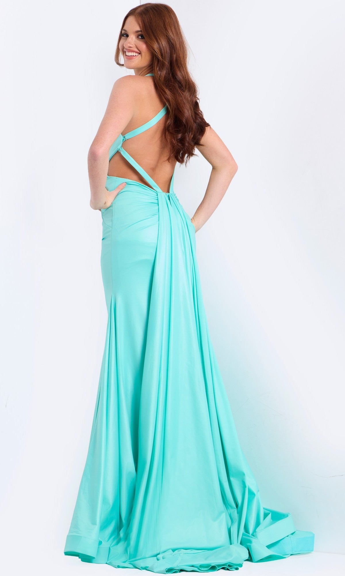 A woman stands smiling with her hands on her hips, showing off the strappy open back of the Jovani 48291 long prom dress in light blue, facing away from the camera against a plain white background.